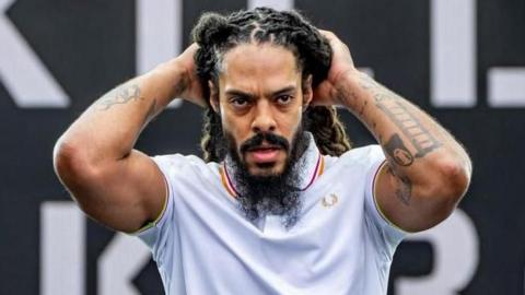 A man performing on stage during a Bob Vylan set.  He is wearing a white polo shirt and his hands are touching his head. He has a dark beard and dreadlocks and tattoos on both his arms.