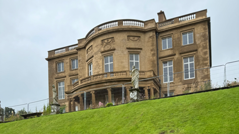 An 18th Century mansion in the Palladian style sits on top of a grassy hill with its facade surrounded by security fencing