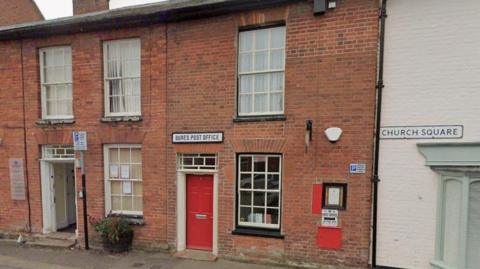A red brick building with a sign saying Bures Post Office. It has a red door and two windows. There is a post box on the outside of the building. There are other buildings either side of the post office.