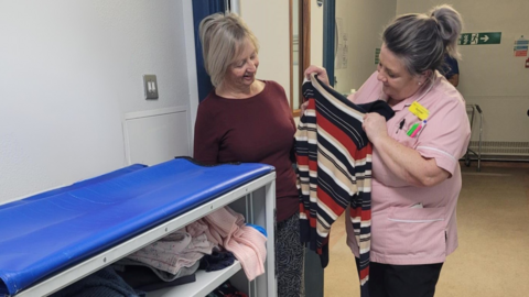 Two women in a hospital. One woman has short blonde hair and is wearing a burgundy top. She is looking at a woman beside her. The other woman has silver/purple hair and is wearing a pink nurse's top. She is holding up a stiped top and showing it to the other woman. Beneath them is a cupboard filled with clothes.