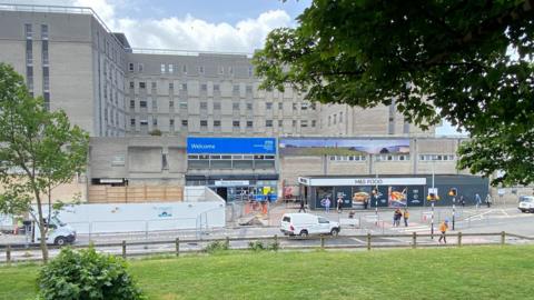 The exterior of Derriford Hospital. There is a large blue sign above the entrance doors that reads WELCOME with NHS branding. There is a single storey M&S Food shop to the left of the entrance. A road runs parallel to the building with a pedestrian crossing on it. In the foreground is a fenced off area of grass with a couple of small trees.