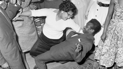 A West Indian couple dancing together at the first Notting Hill Carnival in 1959. Both are wearing traditional 1950s clothes the man is in a suit and the woman is in a white blouse and black skirt. The man is leaning back onto the floor while the woman holds his back while they dance. They are surrounded by other dancers