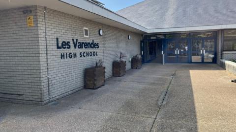 An exterior picture of Les Varendes High School, there is a large sign with the name. The building is a grey brick building with blue rimmed glass doors. 