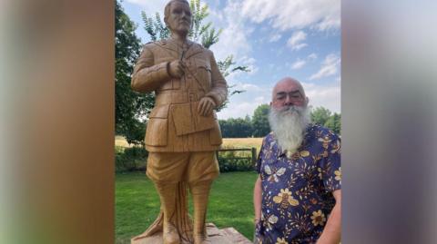 A bearded man in a blue shirt decorated with yellow bees stands next to a wooden statue of JRR Tolkien, that is showing cracks.
