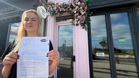 Jennifer Smith holding a letter from Durham County Council telling her she needs to repaint her business' front door. Jennifer Smith Aesthetics has a light pink door with artificial pink roses above it. Jennifer has straight long blonde hair and is wearing bracelets, earrings and a black top.