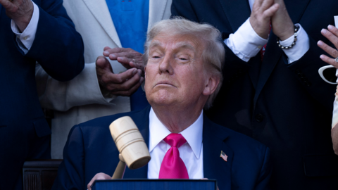 Donald Trump wearing dark suit with a vibrant pink tie, confidently holding up a gave. He is surrounded by people applauding.  This is a photo opp after he signed the "Big Beautiful Bill Act" at the White House in Washington, DC, on July 4, 2025.