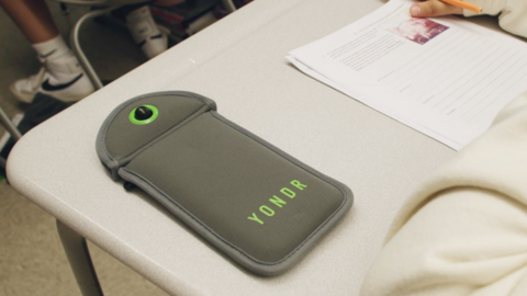 Grey phone pouch with YONDR written on it in green lettering laying on a pupil's desk