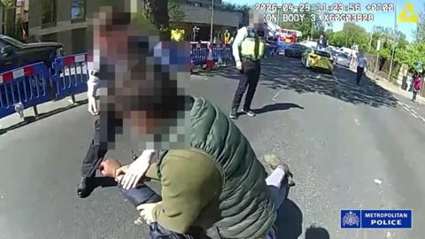 A man with a blurred face wrestles with a man on the ground as police officers stand nearby on a still from police bodycam footage