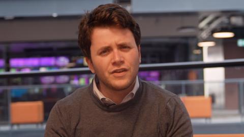 A man looks into the camera, he is wearing a grey jumper, with a blue collared shirt.