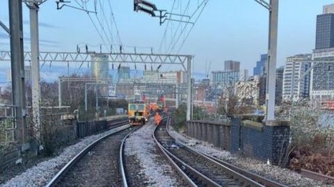 Two sets of rail tracks can be seen going into the distance, where a maintenance train and staff can be seen working. In the background is Birmingham's city skyline. 