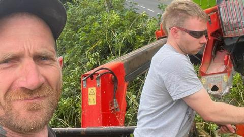 A selfie taken by Daniel Graham shows Daniel Graham in the foreground and Adam Carruthers working behind him. Daniel Graham is looking at the camera and wearing a black cap and black shirt. Adam Carruthers is side on and wearing sunglasses and a grey top