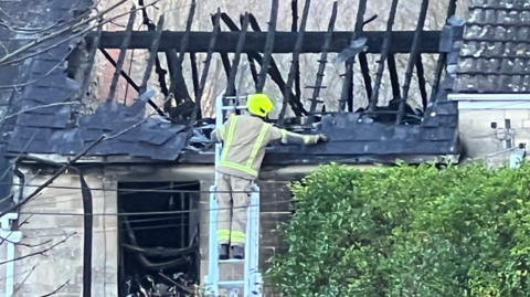 A fire officer is stood on a ladder inspecting the roof of a burned down house. They are wearing reflective gear. The roof is clearly burned through, a blackened room can also be seen through an opening in the brickwork, possibly once a window.