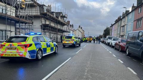 London Road in Chippenham with houses on either side and cars parked. There are three police cars with sirens stopped on the road.