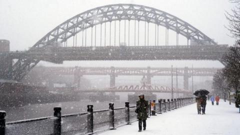 Snow covers the Newcastle Quayside. People walk by the river wearing thick coats and holding umbrellas. The Tyne Bridge, Swing Bridge and High Level Bridge can be seen in the background.