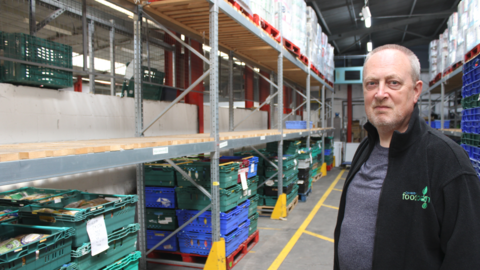 A man with grey hair and a black jacket stands in a warehouse. Rows of empty shelves and stacks of crates can be seen behind him.