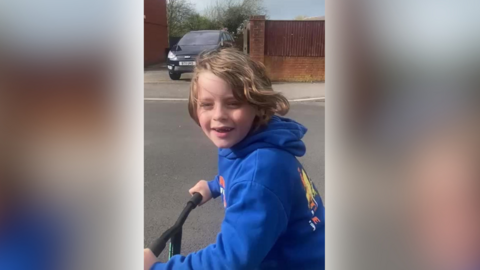 A boy in a blue hoodie on a scooter. He smiles at the camera and his brown hair blows in the wind