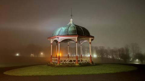 A Victorian-style bandstand stands illuminated in a misty park at night. 