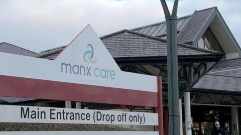 The front of Noble's Hospital, which has a pitched roof and a canopy held up by pillars in front of it. A white sign with the Manx Care logo on it is to the left.