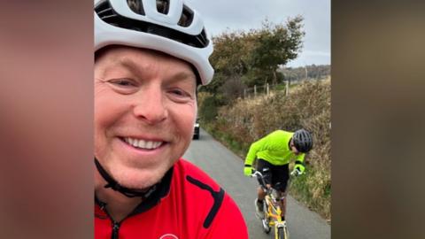 Sir Chris Hoy takes a selfie smiling at the camera with Paddy McGuinness on his bike in the background struggling