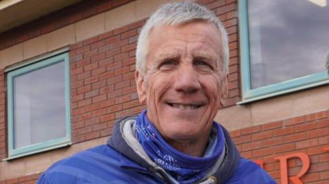 Neil Hunter has short grey hair, is wearing a blue scarf and windbreaker-type jacket, and is smiling for the camera.