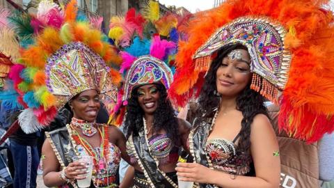 Three women dressed in carnival outfits with bright feathers on top of their heads and two of the women hold drinks