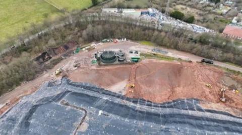 Aerial shot of a muddy-looking hill, partly covered by black sheeting. The site is lined by trees with other buildings behind.