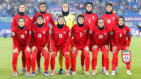 Iran's team pose for a group photo before the AFC Women's Asian Cup Australia 2026 match between Iran and the Philippines in Gold Coast on March 8, 2026. They are wearing red team kit and black hijabs. The goalie wears a yellow top. They are smiling for the camera