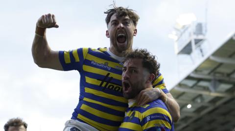 Warrington Wolves' Toby King shouts to celebrate his try while team-mate Matty Ashton leaps on his shoulder and punches the air. Both are wearing Warrington's blue and yellow hoops.