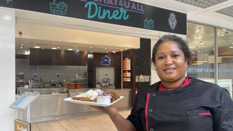 A lady in a black chef's shirt with pink trrim holds a plate of goat curry and rise. She stands in front of a diner inside a shopping centre with a large open entrance and an open kitchen.