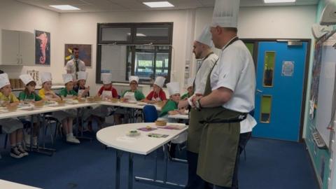 Two professional chefs standing in front of a classroom of children sat down with fresh food and bowls in front of them.