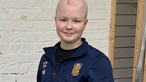 Finn wearing a navy blue cricket tracksuit standing in front of a brick wall painted white. He is smiling.
