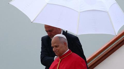 Pope Leo wearing papal cap and robes walks off a plane as a man in a dark suit behind him holds a white parasol over him.