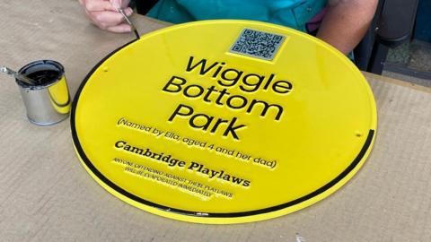 Someone painting a yellow plaque with a black trim. The sign reads 'Wiggle Bottom Park, named by Ella, aged 4 and her dad. Anyone offending against these playlaws will be evaporated immediately'