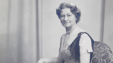 A black and white photo of a lady sat in an arm chair with a long dress on