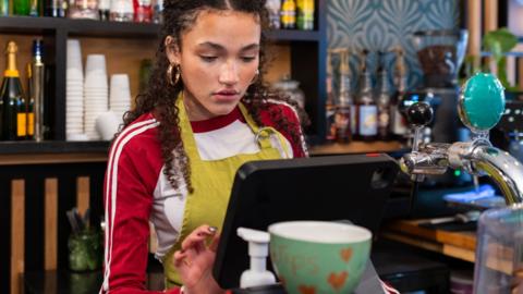 A young woman stands at a till as she works in a cafe and bar