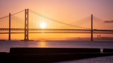 The sun sets over the Forth Road bridge from South Queensferry. The sky is orange and the water is sparkling and dark purple.
