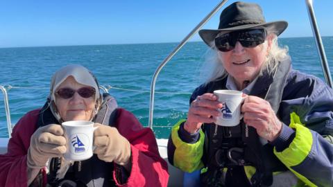 A woman wears a red coat, sunglasses and holds a white Sailing for the Disabled mug. A man sits next to her with long white hair and a hat. He also wears sunglasses and a bright jacket. The blue sea and cloudless skies are behind them on a calm day.
