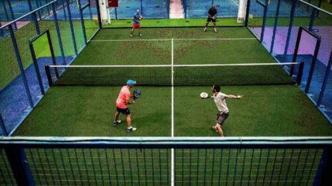 Four people playing a doubles match on an enclosed padel court, with one player in the foreground swinging their racket as the others prepare to return the ball, all inside a brightly lit indoor sports facility.
