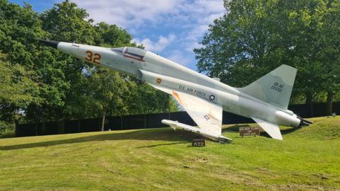 A replica of a F-5 Tiger II is mounted on grass. It is painted blue has U.S AIR FORCE written on the side. There is a fence in the background and several trees.