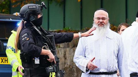 Armed police officer talk to Rabbi Walker in front of a wall with green fencing above