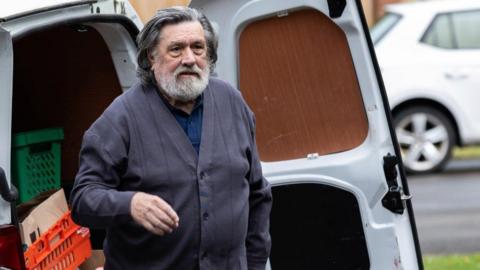 Ricky Tomlinson, who has dark grey hair and a grey beard, is pictured on the set of Brookside. He is standing in front of a white van with one of its back doors open and colourful plastic crates and cardboard boxes visible inside. He is wearing a navy polo shirt and dark grey cardigan. 