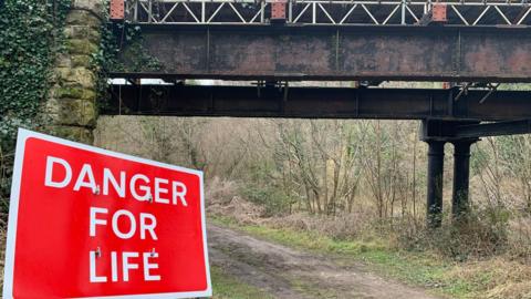 A view of the bridge from the ground below: a large black painted steel structure with some rust, and scaffolding can be seen at the top. Stones at the side. In the foreground of the picture is a large red temporary official sign saying Danger For Life.