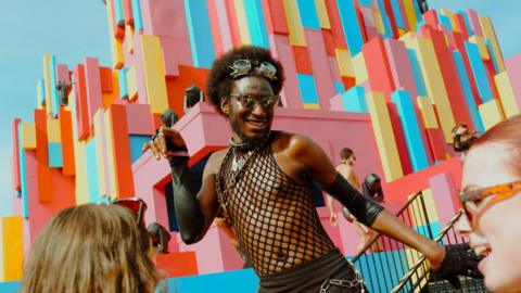 A person wearing a black fishnet string vest and dark bottoms with a silver chain around the waist. They are wearing silver goggles on their head, sweeping back their dark curly hair. Behind them is a brightly coloured brick-style structure that people are walking in.