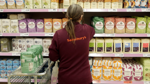 Female Sainsbury's worker in maroon jacket with orange logo on the back stacks bags of flour