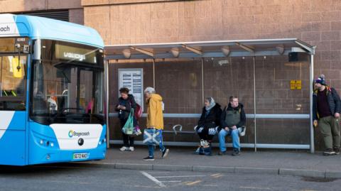A blue and white Stagecoach bus pulls up at a bus stop in Inverness city centre. A woman wearing a yellow coat walks past the bus shelter where a woman is standing and a man and woman are sitting on a bench. A man wearing a woolly hat with a pom-pom leans against the bus shelter.