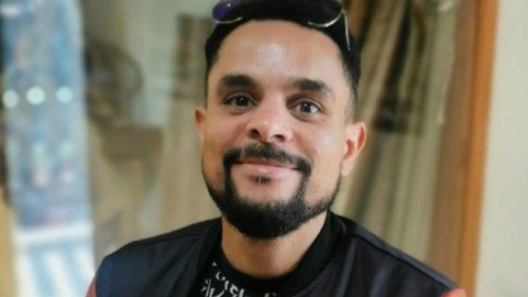A man with black hair and facial hair. He has sunglasses on his head and is wearing a black and white t-shirt and a leather bomber jacket that is black with orange sleeves and patches all over it.
