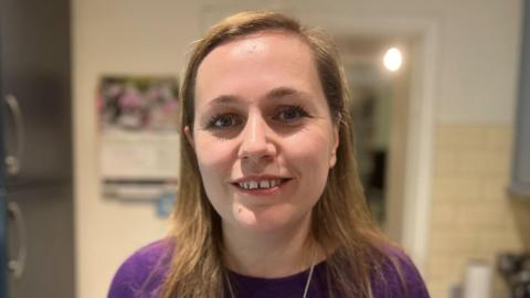 Jen Craft standing inside a kitchen, which is blurred in the background, and smiling. She has long fair hair and is wearing a purple top.