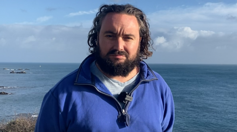 Lee van Katwyk is looking serious as he stands with the sea behind him on a bright blue sky day. He has shoulder-length dark curly hair and a dark beard and moustache. He is wearing a blue half-zipped sweatshirt over a pale blue top and has a microphone clipped to his collar. 