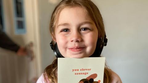 Esmai smiles at the camera while holding a beige card with a photo of a sausage dog on. It reads 'you clever sausage'. She is wearing earphones and a pink top. She has blue eyes and long dark blonde hair. 