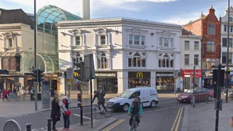 A McDonald's shop front at a busy junction with a small van and two cars, two cyclists and pedestrians at a pelican crossing.